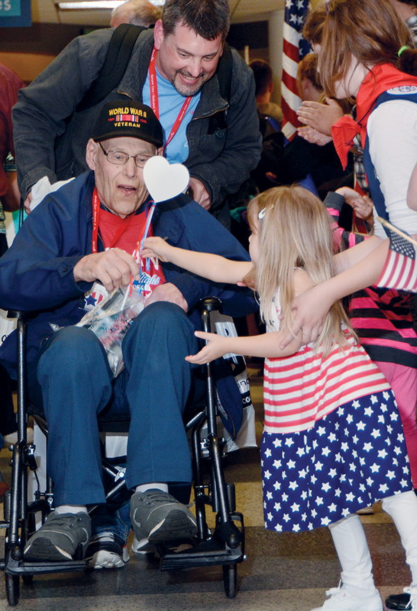 10:09 pm: In a scene repeated from the day’s beginning, veteran Charles Krause is handed a red, white and blue heart from one of many children greeting the returning flight. His guardian, grandson Marty Krause Jr., looks on.
