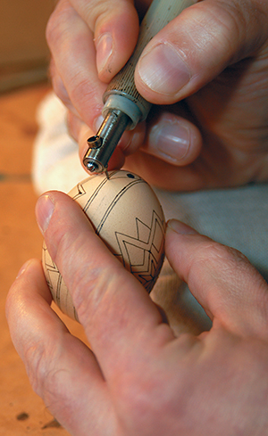 Batz uses an electric “kistka” to draw lines of beeswax, mixed with a black crayon, onto an egg. Beeswax is scooped into the tiny opening at the top, is melted, and comes out of the tip.
