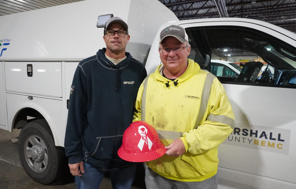 NIPSCO Lineman Aaron Brenneman (left) stands with Marshall County REMC Lineman John Yates (right), showcasing a custom pink hard hat that Brenneman made for Yates.