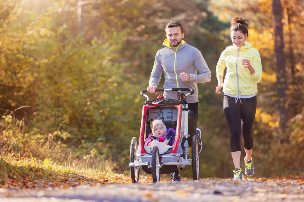 Beautiful young family with baby in jogging stroller running outside in autumn nature